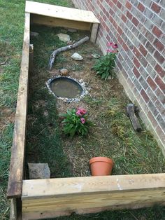 an empty wooden box sitting in the grass next to a brick wall and potted plants