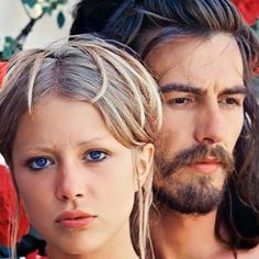 two people with long hair and blue eyes standing next to each other in front of flowers