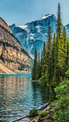 the mountains are covered in snow and pine trees near a lake with clear blue water