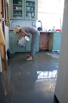 a woman in grey shirt pouring water into a white bucket on top of a kitchen floor