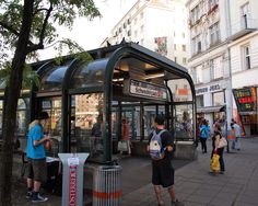 people are standing in front of a bus stop on the sidewalk near tall buildings and trees