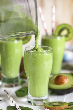 a green smoothie being poured into a glass with avocado slices around it