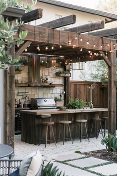 an outdoor kitchen and dining area with lights strung from the ceiling, surrounded by greenery