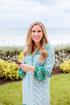 a woman standing in the grass holding a cell phone and smiling at the camera with her hand on her hip