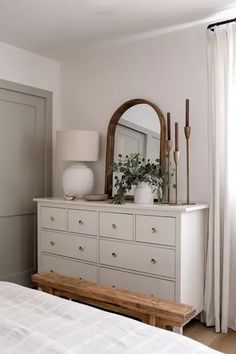 a bedroom with a dresser, mirror and lamp on it's side table in front of a window