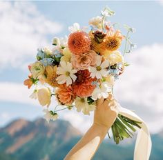 a person holding a bouquet of flowers in front of a mountain range with blue sky and clouds