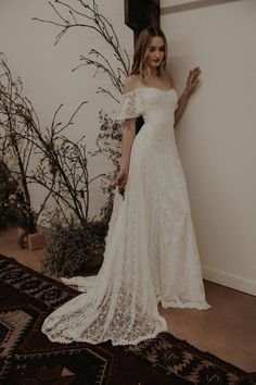 a woman standing in front of a wall wearing a white wedding dress with off the shoulder sleeves