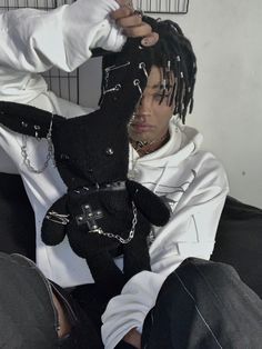 a man with dreadlocks is holding a black teddy bear in his lap and wearing a white shirt