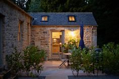 a small stone house lit up at night with lights on the windows and patio furniture