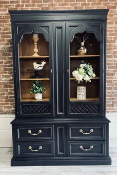 a black china cabinet with flowers and vases on it in front of a brick wall