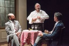 three men sitting at a table with wine glasses in front of them and one man standing up