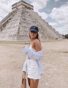 a woman is standing in front of an ancient pyramid wearing a hat and holding a straw bag