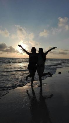 two women standing on the beach with their arms in the air as the sun sets