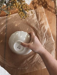 a person holding a coffee cup on top of a wooden table next to dried flowers