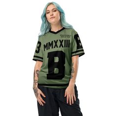 a woman with blue hair wearing a green football jersey and black pants, standing in front of a white background