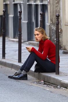 a woman sitting on the curb looking at her cell phone while wearing a red sweater and black pants