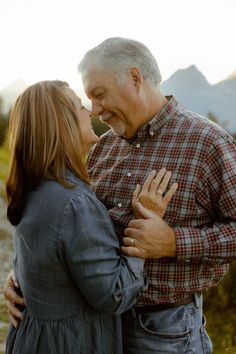 an older man and woman kissing each other in front of the camera with mountains behind them