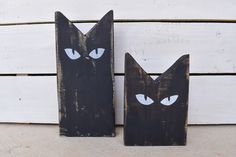 two black wooden cats with blue eyes are sitting on a white wood planked wall