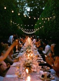 a long table with people sitting at it and lit up by string lights in the background