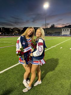 two cheerleaders pose for a photo on the football field at night with flowers in their hands