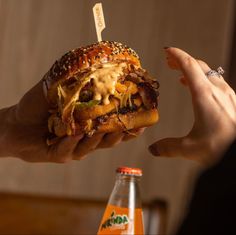 a person holding up a large sandwich with cheese and other toppings next to a bottle of soda