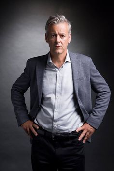 a man with his hands on his hips wearing a suit and white shirt, standing in front of a dark background