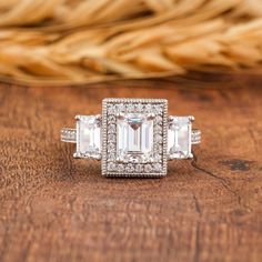 an engagement ring with three emeralds and diamonds on a wooden table next to some wheat