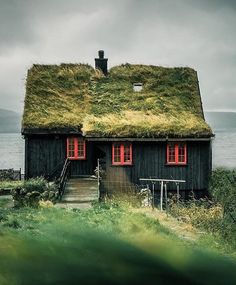 an old black house with grass on the roof and red windows, next to water