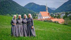 three women standing in front of a church on top of a lush green hillside with mountains in the background