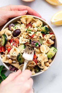 two hands holding a fork over a bowl of pasta salad with cucumbers, tomatoes and olives