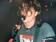 a man with long hair playing guitar in front of a microphone and another person standing behind him