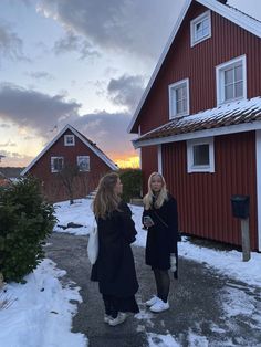 two women standing in front of a red house with snow on the ground and trees