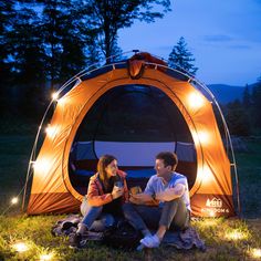 a man and woman sitting in front of an orange tent with lights on it at night