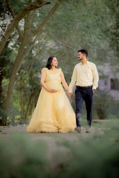 a pregnant woman in a yellow dress and a man in a white shirt hold hands as they walk through the woods