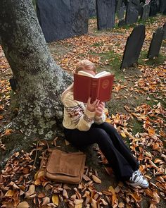 a woman sitting on the ground reading a book next to a tree and tombstones