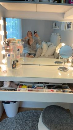 a woman taking a selfie in front of a white vanity with drawers and lights