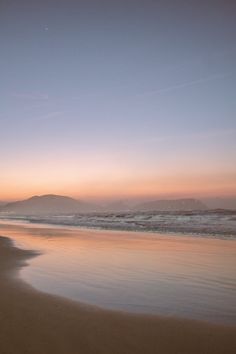 two people walking on the beach at sunset with surfboards in hand and mountains in the background