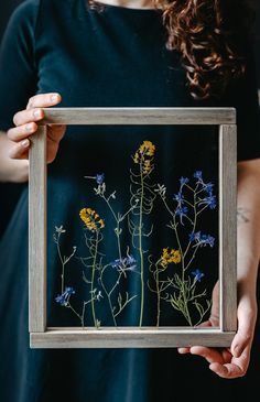 a woman holding up a framed painting with wildflowers in the frame on it