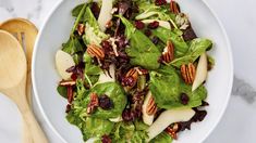 a salad with apples, cranberries and pecans in a white bowl next to a wooden spoon