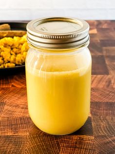 a jar filled with yellow liquid sitting on top of a wooden table next to a tray of food