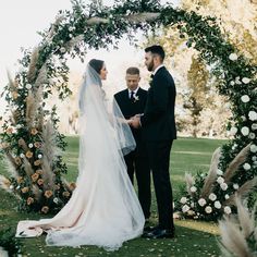 a bride and groom are standing under an arch with flowers on the grass in front of them