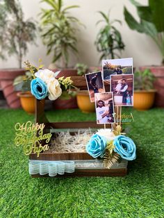 an open wooden box with photos and flowers in it sitting on the grass next to potted plants