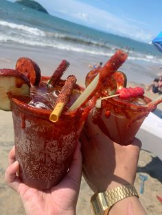 two people holding up glasses with food on the beach and water in the ocean behind them