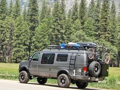 a truck with bikes on the roof parked in front of some pine trees and mountains