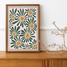 an art print with green and yellow flowers on a white wall next to a wooden dresser