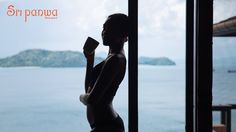 a woman standing in front of a window drinking out of a cup next to the ocean