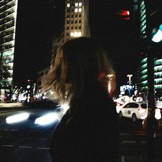 a woman standing on the side of a road at night with traffic lights in the background