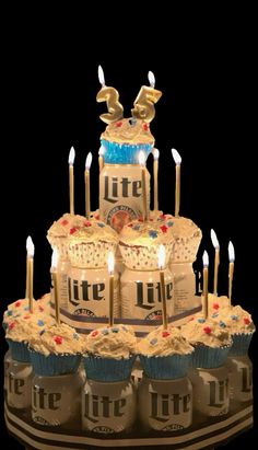 a birthday cake with lit candles in the shape of two beer cans and cupcakes