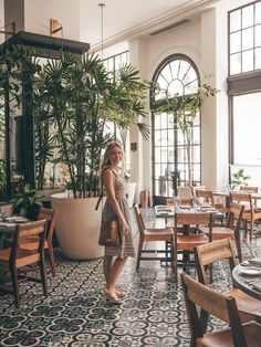 a woman is standing in the middle of a restaurant with tables and chairs, potted plants behind her