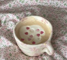 a white cup with hearts on it sitting on a pink flowered cloth covered tablecloth